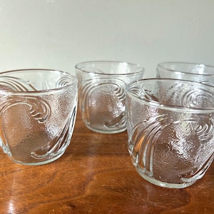May include: Four clear, textured glass tumblers with an embossed swirl design. The glasses are arranged on a wooden surface, showcasing their decorative pattern. The glasses are suitable for serving beverages.