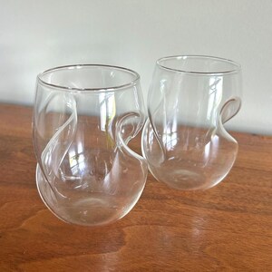 May include: Two clear, stemless wine glasses with a distinctive, swirling design. Each glass has a curved handle-like feature that wraps around the body. The glasses are made of transparent glass and sit on a wooden surface.