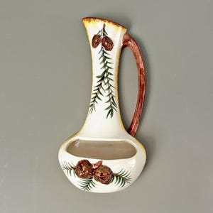 May include: White ceramic wall planter shaped like a pitcher with a brown handle. The planter is decorated with green pine branches and brown pine cones.