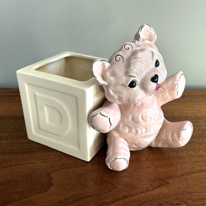 May include: A pink ceramic teddy bear planter with a white ceramic block planter shaped like a letter D. The teddy bear planter has black eyes and a black nose.