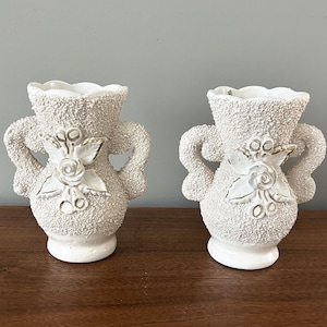 May include: Two white ceramic vases with gold accents and a raised floral design. The vases have two handles on each side.