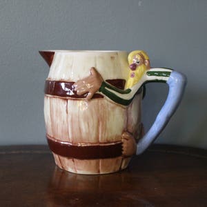 May include: A ceramic pitcher shaped like a barrel with a cartoon man holding onto the handle. The barrel is painted to look like wood with brown bands. The man has a yellow beard, blue shirt, and green and white striped sleeves.