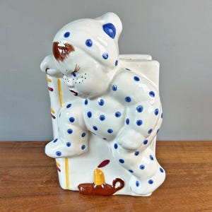 May include: A vintage ceramic figurine of a baby in pajamas with blue polka dots, leaning against a white rectangular form. The baby has brown hair and is next to a candle. The figurine is on a wooden surface.