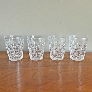 May include: Four clear glass tumblers arranged in a row, each featuring a repeating pattern of raised circular bubbles across the exterior surface. The glasses have a straight-sided cylindrical shape with a smooth rim and a thick, solid base.