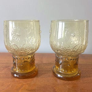 May include: Two amber-colored vintage drinking glasses. The glasses have a floral design etched into the glass. The base of the glasses is a darker amber color with a decorative pattern. The glasses are on a wooden surface.