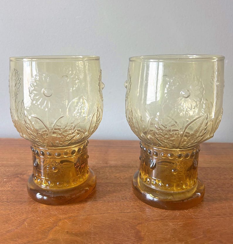 May include: Two amber-colored vintage drinking glasses. The glasses have a floral design etched into the glass. The base of the glasses is a darker amber color with a decorative pattern. The glasses are on a wooden surface.