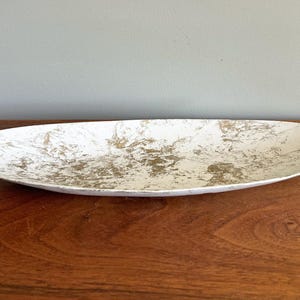 May include: An oval-shaped shallow dish featuring a white base color with an irregular, mottled gold-toned metallic pattern across the interior surface. The dish has a smooth, curved rim and a wide, elongated profile with a low-profile design.