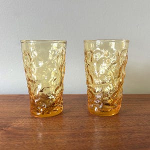 May include: Two amber-colored drinking glasses with a textured, organic pattern. The glasses are cylindrical and sit on a wooden surface. The textured design gives the glasses a unique, vintage look.