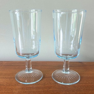 May include: Two clear, stemmed glasses with a light blue tint. The glasses have a flared bowl shape and a round base with a sunburst design. The stems are slender, and the rims are also light blue.