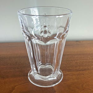 May include: A clear, faceted glass with a hexagonal base and a slightly flared top. The glass features a decorative bee design near the top and vertical ridges along its sides. The glass is empty and sits on a wooden surface.