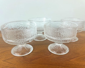 May include: A set of four clear glass dessert bowls with textured, bark-like exteriors. The bowls have a rounded shape and sit on a short, stemmed base. The glassware is arranged on a wooden surface.