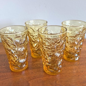 May include: A set of four amber-colored drinking glasses featuring a textured, organic, and uneven surface pattern. Each glass has a cylindrical shape with a slightly flared rim and a thick base, showcasing a translucent, warm yellow-orange hue.