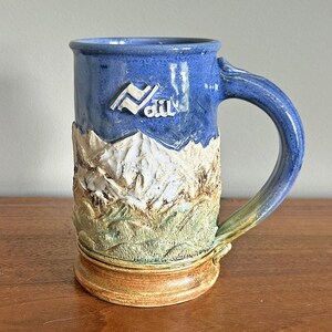 May include: A tall ceramic mug featuring a blue upper section with the embossed white text Vail, a central relief of snow-capped mountains, a green textured landscape base, and a sturdy curved handle with a blue and tan glazed finish.