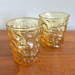 May include: Two amber-colored glass tumblers featuring a textured, organic, and uneven surface pattern. The glasses have a wide, open rim and a slightly tapered base, showcasing a translucent, warm yellow hue that catches the light across the surface.