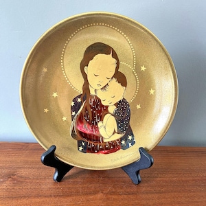 May include: A gold-colored ceramic plate with a painting of a mother and child. The mother is holding the child close to her, and they are both surrounded by stars. The plate is on a black stand.