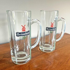 May include: Two clear glass beer mugs with handles. Each mug has the Heineken logo in black, white, and red. The mugs are on a wooden surface.