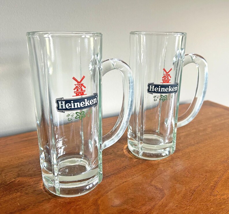 May include: Two clear glass beer mugs with handles. Each mug has the Heineken logo in black, white, and red. The mugs are on a wooden surface.