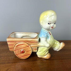 May include: A ceramic planter shaped like a boy pushing a wheelbarrow. The boy has blonde hair, blue eyes, and is wearing a blue shirt and yellow pants. The wheelbarrow is brown with red stripes and a red wheel.