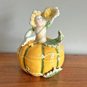 May include: A ceramic pumpkin-shaped container with a lid, featuring a smiling figure emerging from the top. The pumpkin is yellow with green stripes and vines. The figure has a sunflower head and holds a flower. A unique decorative piece.