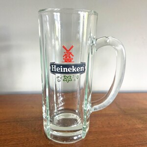 May include: A clear glass beer mug with a handle. The mug features the Heineken logo, a red windmill and the brand name in white on a black background. The mug is rectangular with vertical lines.