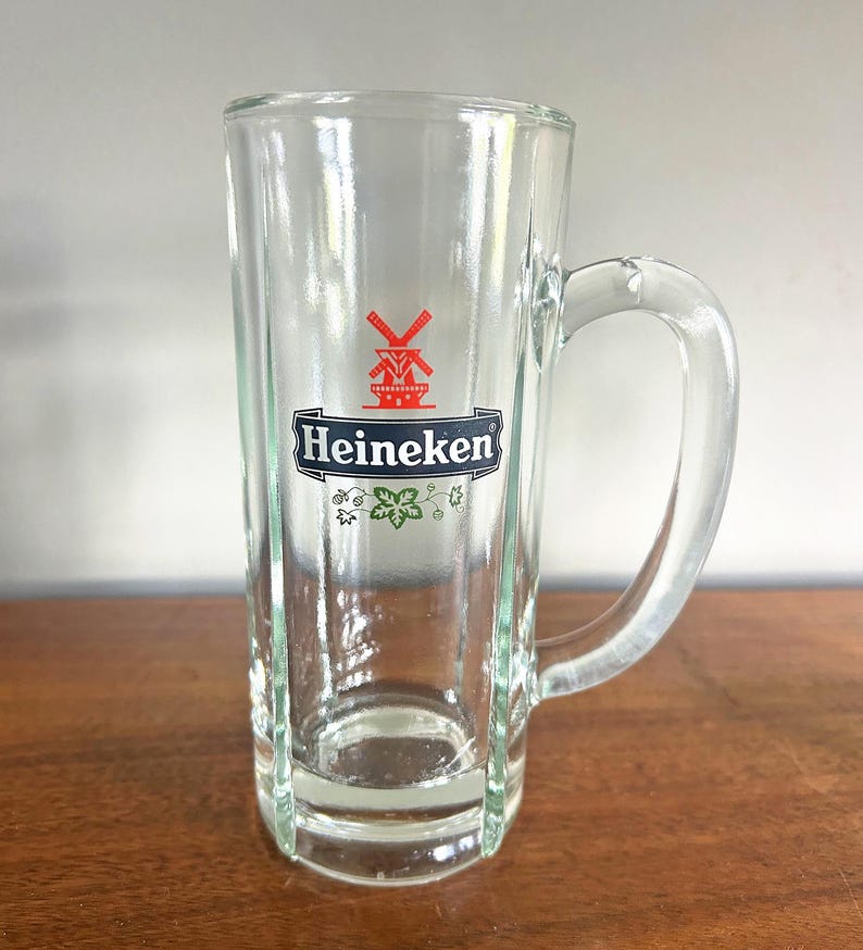May include: A clear glass beer mug with a handle. The mug features the Heineken logo, a red windmill and the brand name in white on a black background. The mug is rectangular with vertical lines.