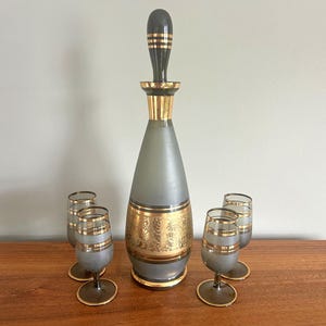 May include: A vintage gray glass decanter set with gold accents. The decanter has a black and gold stopper and is surrounded by four matching glasses. The set features gold bands and floral designs, creating an elegant aesthetic.