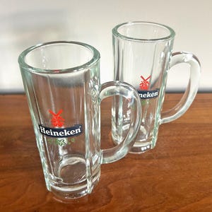 May include: Two clear glass beer mugs with handles. Each mug has a rectangular shape with vertical ridges and a logo that says "Heineken" with a red star and green accents. The mugs are on a brown wooden surface.