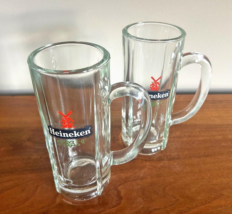 May include: Two clear glass beer mugs with handles. Each mug has a rectangular shape with vertical ridges and a logo that says "Heineken" with a red star and green accents. The mugs are on a brown wooden surface.