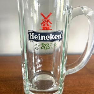 May include: Clear glass beer mug with a handle. The mug features the Heineken logo in red and white, with a windmill graphic. The mug has a rectangular shape with vertical grooves and a curved handle.