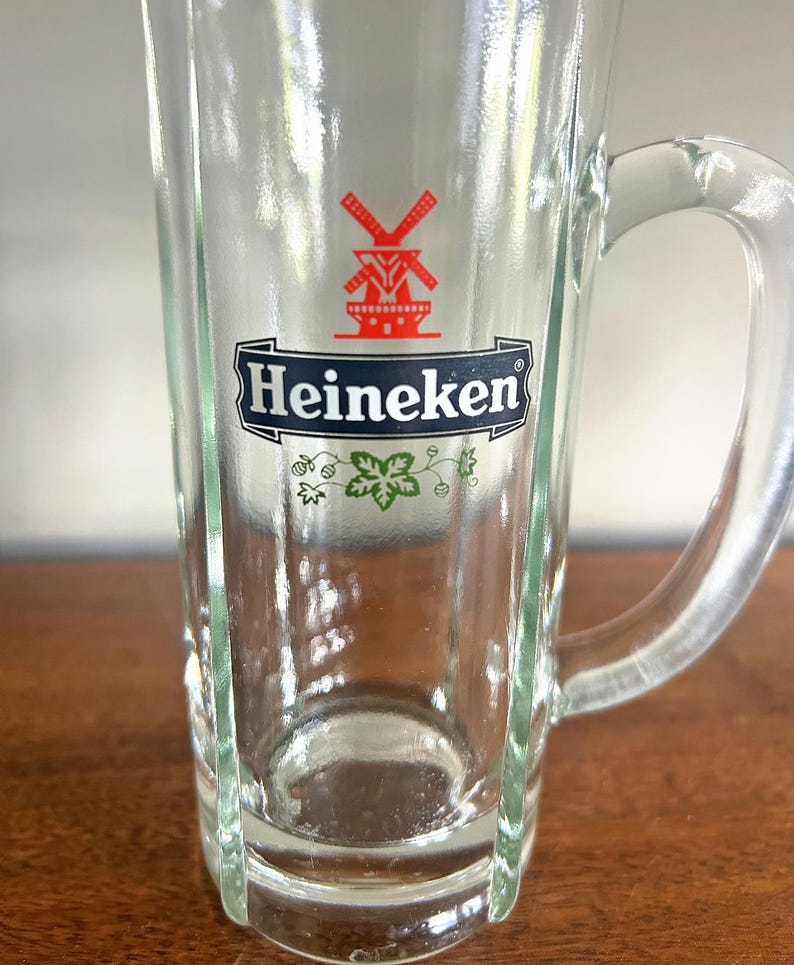 May include: Clear glass beer mug with a handle. The mug features the Heineken logo in red and white, with a windmill graphic. The mug has a rectangular shape with vertical grooves and a curved handle.