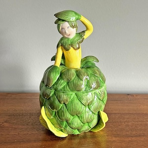May include: A whimsical ceramic artichoke-shaped container with a lid featuring a figure. The figure has a yellow torso and arms, green leafy collar and hat. The artichoke is green with yellow accents.