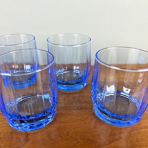 May include: Four clear drinking glasses with a vibrant blue base and rim. The glasses have a textured, ridged design on the lower half. The glasses are empty and sit on a wooden surface.