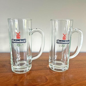 May include: Two clear glass beer mugs with handles. Each mug has the Heineken logo, featuring a red windmill and the brand name in white on a black banner. The mugs are on a wooden surface.
