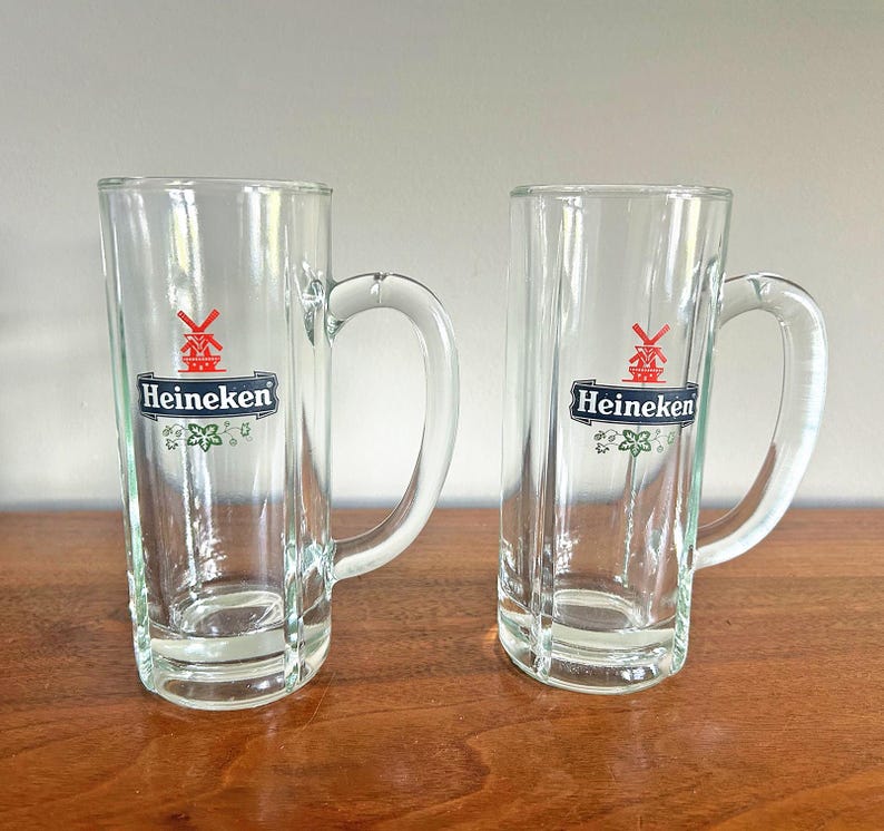 May include: Two clear glass beer mugs with handles. Each mug has the Heineken logo, featuring a red windmill and the brand name in white on a black banner. The mugs are on a wooden surface.