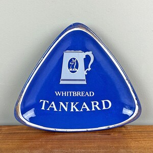 May include: A triangular blue Whitbread Tankard tray with a white border and a gold-colored rim. The tray features a light blue illustration of a tankard and the text "WHITBREAD TANKARD" in white.