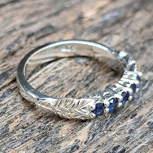 May include: A silver ring with a leaf design on the band. The ring features a row of small, round, deep blue gemstones. The ring is set against a textured, wooden surface.