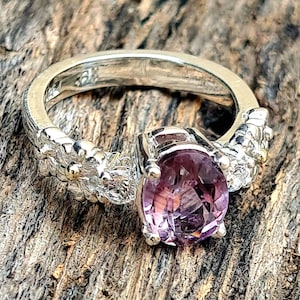 Puede incluir: Anillo de plata con una gran piedra preciosa ovalada de color púrpura. La banda presenta un diseño floral en los laterales. El anillo se encuentra sobre un fondo de madera, resaltando los detalles de la joya.