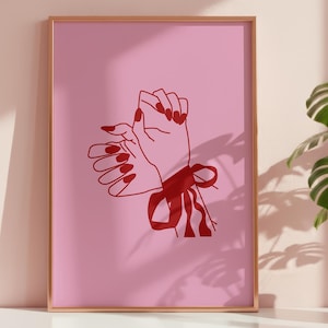 May include: A framed art print. It shows a minimalist illustration of two hands with red nails, tied with a red ribbon, against a pink background. The artwork is in a gold frame.