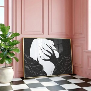 May include: A framed art print featuring a black and white abstract design. The artwork depicts a figure with outstretched hands and surrounding foliage. The frame is gold, and the print is displayed against a pink wall.