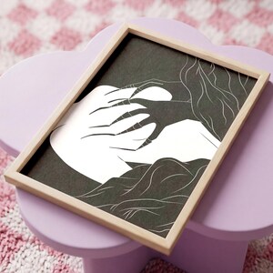 May include: A framed art print featuring a black and white illustration. The artwork depicts a stylized human form with long hair and a hand reaching out. The frame is made of light-colored wood, and the background is a dark color.