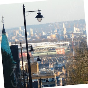 May include: A view of a city skyline with a large stadium in the foreground. The stadium has a white roof and a green field. There are several streetlights in the foreground.