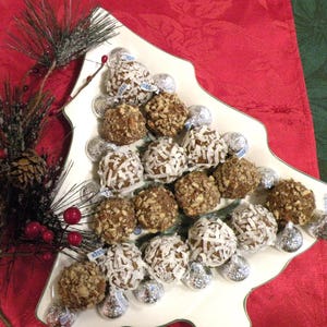 May include: A white ceramic Christmas tree-shaped plate filled with chocolate-covered candies, some topped with shredded coconut. The plate is on a red tablecloth with a green tablecloth in the background.