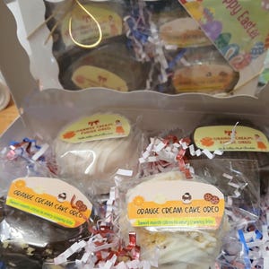 Pu&ograve; includere: Una scatola bianca piena di Oreo alla torta alla crema d'arancia confezionati singolarmente. La confezione include etichette con il testo "Orange Cream Cake Oreo" e "Sweet meets citrus in every creamy bite". La scatola &egrave; decorata con carta sminuzzata rossa, bianca e blu e un'etichetta "Buona Pasqua".