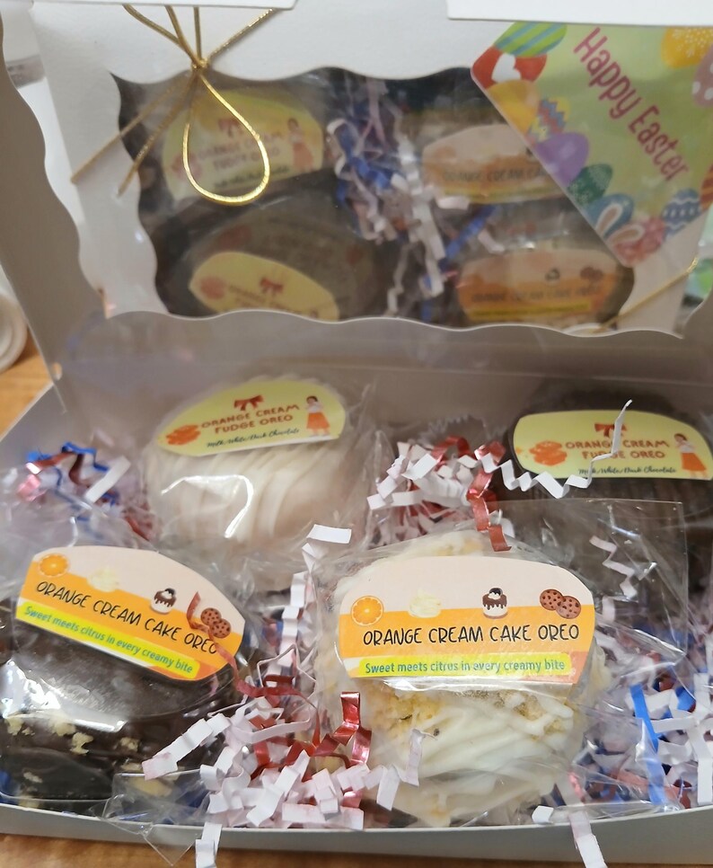 Pu&ograve; includere: Una scatola bianca piena di Oreo alla torta alla crema d'arancia confezionati singolarmente. La confezione include etichette con il testo "Orange Cream Cake Oreo" e "Sweet meets citrus in every creamy bite". La scatola &egrave; decorata con carta sminuzzata rossa, bianca e blu e un'etichetta "Buona Pasqua".