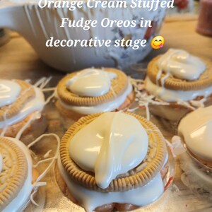 Pu&ograve; includere: Primo piano di diversi Oreo al fudge ripieni di crema all'arancia, ciascuno ricoperto di glassa bianca. I biscotti sono disposti su una superficie decorativa. L'immagine include il testo "Orange Cream Stuffed Fudge Oreos in decorative stage".