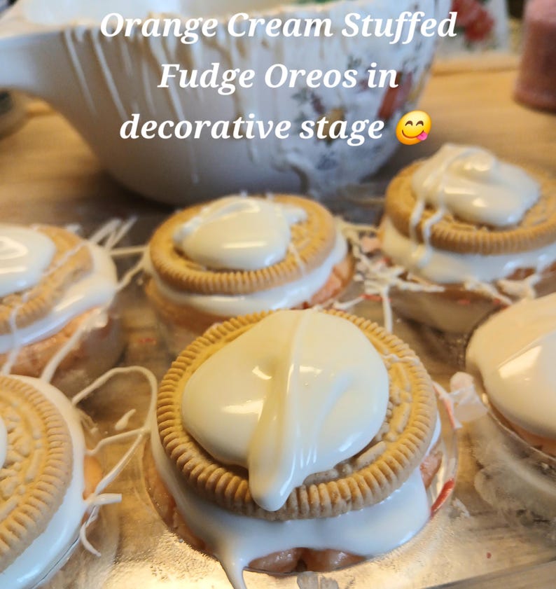 Pu&ograve; includere: Primo piano di diversi Oreo al fudge ripieni di crema all'arancia, ciascuno ricoperto di glassa bianca. I biscotti sono disposti su una superficie decorativa. L'immagine include il testo "Orange Cream Stuffed Fudge Oreos in decorative stage".