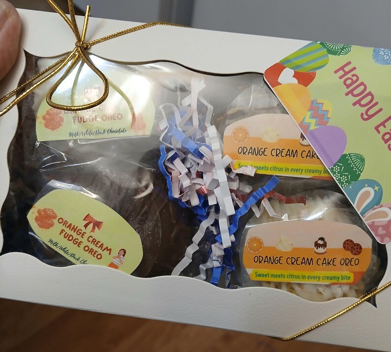 Pu&ograve; includere: Una confezione regalo bianca con una finestra trasparente, contenente biscotti Oreo e torta. I biscotti sono etichettati "Orange Cream Fudge Oreo" e "Fudge Oreo". &Egrave; visibile una carta con "Buona Pasqua".