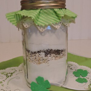 May include: A glass jar with a green and white checkered fabric lid, filled with layers of white powder, brown flakes, and white and brown oats. The jar is sitting on a white doily with green shamrocks.