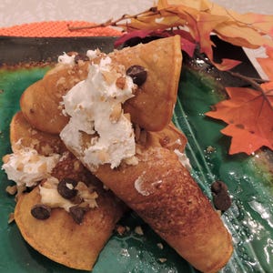 Könnte beinhalten: Ein Stapel Kürbispfannkuchen, belegt mit Schlagsahne, Schokoladenstückchen und gehackten Nüssen. Die Pfannkuchen liegen auf einem grün-braunen Teller mit Herbstblättern im Hintergrund.