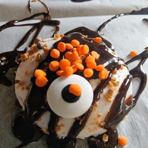 May include: A white chocolate-covered marshmallow treat with a chocolate eye, orange sprinkles, and a chocolate drizzle on top.